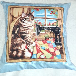May include: A blue pillow with a brown frame featuring a cat sitting next to a vintage sewing machine and a colorful patchwork quilt. The frame has the word "Sewing" on the machine.