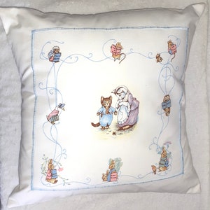 May include: A white pillow with a blue border and a colorful illustration of a cat and a rabbit. The illustration is surrounded by a blue and white floral design. The text "Peter Rabbit" appears on the pillow.