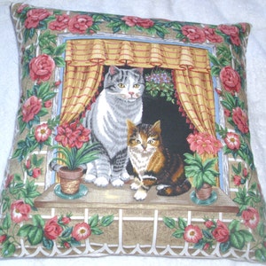 Könnte beinhalten: Ein Dekokissen mit einem Blumendesign, das zwei Katzen zeigt, die aus einem Fenster schauen. Die Katzen sind grau und getigert, und das Fenster ist mit einem weißen Gitter gerahmt. Das Kissen ist beige mit einem weißen Rand.