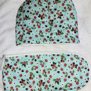 May include: A quilted tea cozy and oven mitt set. The set features a light blue background with a pattern of red raspberries, white flowers, and green leaves. The edges of the cozy and mitt are trimmed with white fabric.
