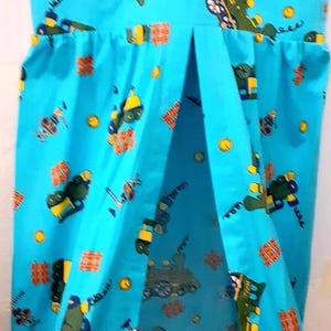 May include: A light blue fabric item with a repeating pattern of green and yellow vehicles, orange blocks, and yellow circles. The item has a gathered top and a split front, revealing a white interior.
