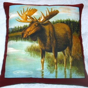 May include: A brown and tan moose with large antlers stands in a lake with green reeds. The moose is looking to the left of the image. The background is a blue sky with white clouds. The image is on a red pillow.