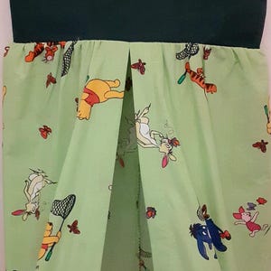 May include: A green fabric hanging organizer with a Winnie the Pooh and friends print. The organizer has a green fabric top and a white fabric bottom. It hangs from a metal hook.