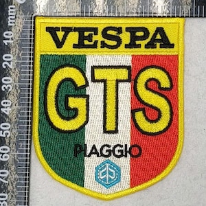 May include: A shield-shaped embroidered patch featuring the text VESPA in black at the top, followed by large yellow GTS letters over a green, white, and red vertical striped background. The word PIAGGIO and a light blue logo are visible below.
