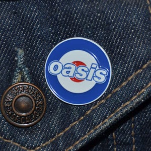 Può includere: Uno spillo in smalto blu e bianco con un cerchio rosso al centro e la parola "oasis" in lettere bianche.