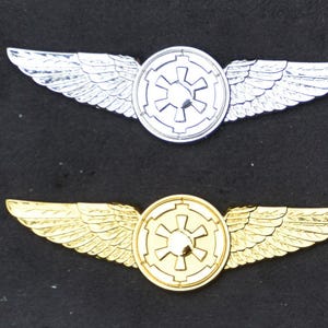 May include: Two metal pilot wings with a circular emblem in the center. The top wings are silver and the bottom wings are gold. The emblem features a stylized star with four points.