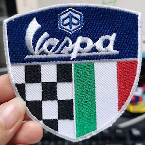 May include: A shield-shaped embroidered patch featuring the word Vespa in white script on a dark blue background. Below, the design is divided into a black and white checkered pattern on the left and the green, white, and red Italian flag on the right.