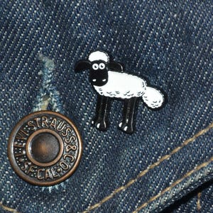 May include: A cartoon sheep pin in black and white, attached to a blue denim jacket. The pin shows a sheep with a white body, black legs, and a black face. A copper button with the text "LEVI STRAUSS & CO. CAL." is also visible.