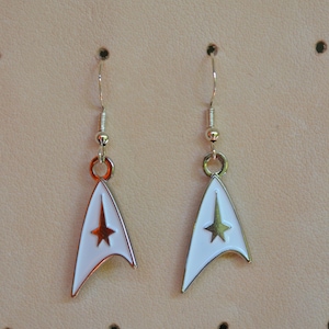 Op de afbeelding: Set van witte en zilverkleurige Star Trek-oorbellen. Elke oorbel heeft een wit geëmailleerd embleem met een gouden ster en een zilverkleurige rand. De oorbellen hangen aan zilverkleurige oorhaken.