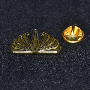 May include: Gold-coloured pin with a stylised design of a fan or a crown. The pin has a simple, round back.