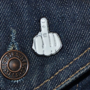 May include: A white enamel pin of a middle finger on a blue denim jacket.