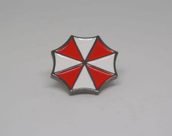 May include: A metal pin in the shape of an umbrella, featuring alternating red and white segments. The pin has a silver-colored border and a glossy finish, set against a plain background.