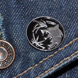 May include: A silver metal pin with a black and white design of a wolf and a bird. The pin is on a blue denim background.