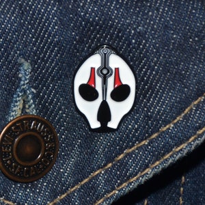 Puede incluir: Pin de esmalte blanco con un diseño negro y rojo de una cara estilizada con forma de calavera. El pin está en una chaqueta vaquera azul.