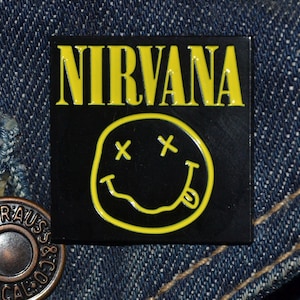 Peut inclure: Épinglette carrée noire avec le mot "NIRVANA" en lettres jaunes au-dessus d'un smiley jaune avec des yeux barrés et une langue tirée. Sur fond denim.