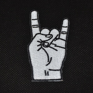 Può includere: Una toppa ricamata bianca di una mano che fa il gesto rock and roll. La toppa è su uno sfondo nero.