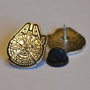 Puede incluir: Un pin de metal plateado con el Halcón Milenario de Star Wars. El pin tiene un respaldo de goma negra y un cierre de metal plateado.