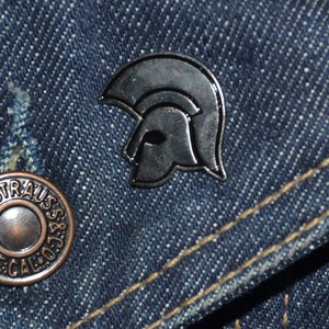 Pode incluir: Um alfinete de metal prateado em forma de capacete com contorno preto. O alfinete está em um fundo de jeans azul.