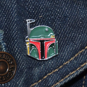 Può includere: Uno spillo in metallo argentato con un design in smalto verde, rosso e nero di un casco di Boba Fett di Star Wars.