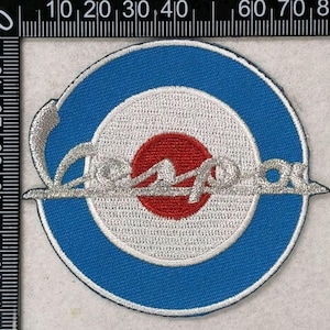 Puede incluir: Un parche bordado circular con un anillo exterior azul, un anillo central blanco y un círculo rojo en el centro. El texto "Vespa" está bordado en color plateado sobre el diseño. Una regla muestra que el parche mide unos 8,2 cm de ancho.