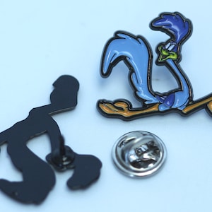 May include: A blue and yellow cartoon roadrunner enamel pin with a black outline. The pin is attached to a silver metal pin back.