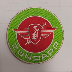 May include: A round green and red embroidered patch with a white winged shield and the text "NUNDAPP" in white.