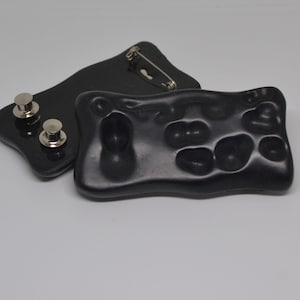 May include: Two black resin brooches, one with a textured, abstract design and the other with a simple pin closure.