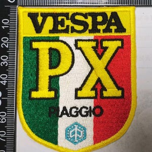 May include: A shield-shaped embroidered patch featuring the text VESPA in black, large yellow PX letters, and the word PIAGGIO in black. The background displays vertical green, white, and red stripes with a small blue hexagonal logo at the bottom.