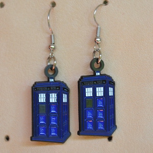 May include: A pair of blue and black earrings shaped like the iconic British police call box, the TARDIS. The earrings have silver-colored hooks and black circular connectors. The TARDIS design features white windows and a black outline.