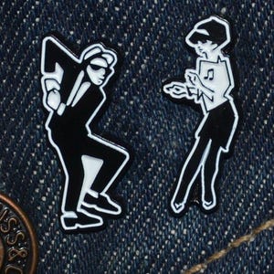 Op de afbeelding: Twee zwart-witte emaille pins met gestileerde dansende figuren. Eén figuur is een man in pak en zonnebril, de andere een vrouw met bladmuziek. De pins zijn geplaatst tegen een blauwe denim achtergrond.