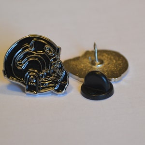 Könnte beinhalten: Eine schwarze und silberne Emaille-Anstecknadel mit einem detaillierten Design eines Helms. Die Nadel hat eine silberne Rückseite mit einem schwarzen Gummifixierer.