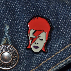Könnte beinhalten: Emaille-Anstecknadel mit einem stilisierten Porträt mit rotem Haar, weißem Gesicht und einem Blitz auf dem Gesicht. Die Anstecknadel hat einen schwarzen Rand, vor einem blauen Jeans-Hintergrund.