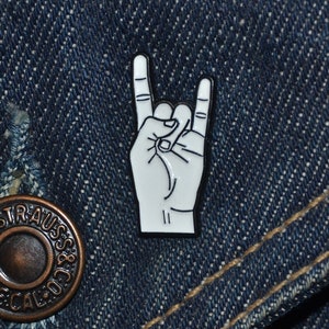 Może przedstawiać: Biała emaliowana przypinka z ręką wykonującą gest rock and roll. Przypinka jest na niebieskiej dżinsowej kurtce.