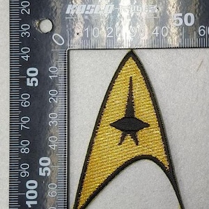 Puede incluir: Parche bordado dorado y negro con la insignia de Starfleet. El parche tiene forma de triángulo con un diseño de estrella y flecha negra. El parche mide aproximadamente 6,3 cm de alto.