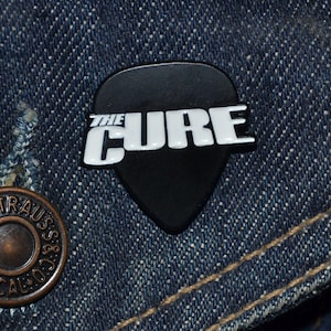Op de afbeelding: Zwarte gitaarplectrumvormige pin met witte tekst "THE CURE" op een denim achtergrond.