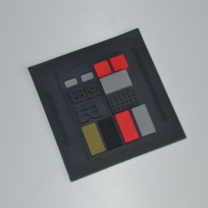 Könnte beinhalten: Ein schwarzes Star Trek Kommunikatorabzeichen aus Gummi mit einem erhabenen Design aus Knöpfen und einem Bildschirm. Das Abzeichen trägt den Text "www.woodlesticks.com" in der unteren rechten Ecke.