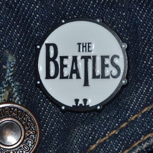 Puede incluir: Un pin de esmalte blanco y negro con un tambor que tiene el texto "The Beatles" en él.