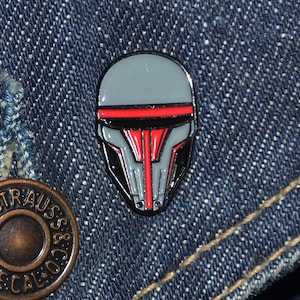 Może przedstawiać: Szpilka emaliowana w kolorze szarym i czerwonym przedstawiająca stylizowany hełm. Hełm ma czerwony pasek na górze i czarną osłonę.