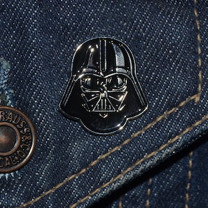 Könnte beinhalten: Eine silberfarbene Darth Vader-Anstecknadel mit einem schwarz-weißen Design. Die Anstecknadel befindet sich auf einer blauen Jeansjacke.