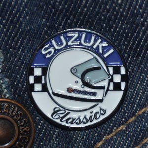 May include: A white and blue enamel pin with a checkered flag design. The pin features a white motorcycle helmet with a Suzuki logo and the text "Suzuki Classics".