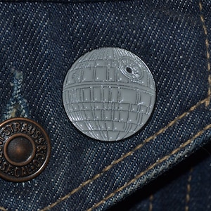 May include: A white enamel pin shaped like the Death Star from Star Wars. The pin is on a blue denim jacket.