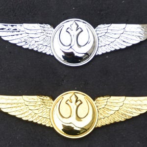 Könnte beinhalten: Zwei Rebellen-Allianz-Pilotenflügel, einer silber und einer gold, mit dem Symbol der Rebellen-Allianz in der Mitte. Die Flügel sind aus Metall und haben ein detailliertes Design.