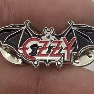 Könnte beinhalten: Metall-Anstecknadel mit schwarzer Fledermaus und roter und weißer Aufschrift "OZZY". Die Nadel hat zwei Nadelrücken.