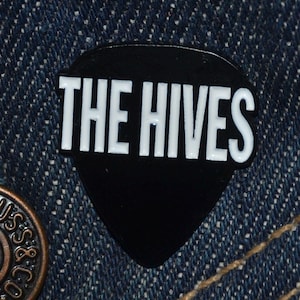 Puede incluir: Un pin negro con forma de púa de guitarra con el texto blanco "THE HIVES" sobre un fondo de mezclilla. El pin tiene un acabado brillante. El texto está en negrita, con una fuente sans-serif.