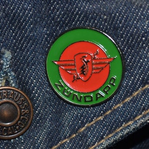 May include: A green, red, and black enamel pin with the text "Nundapp" and a stylized winged shield design.