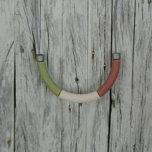 Op de afbeelding: Een ketting met een gebogen, veelkleurige houten hanger. De hanger is gemaakt van drie delen: groen, wit en rood. De hanger hangt tegen een witgeschilderde houten achtergrond.