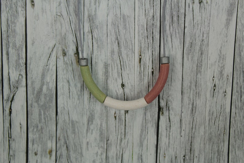 Op de afbeelding: Een ketting met een gebogen, veelkleurige houten hanger. De hanger is gemaakt van drie delen: groen, wit en rood. De hanger hangt tegen een witgeschilderde houten achtergrond.