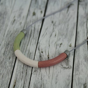 Op de afbeelding: Een ketting met een enkele, grote, veelkleurige kraal. De kraal is gemaakt van drie delen: groen, wit en roze. De ketting is gemaakt van een dun, grijs koord.
