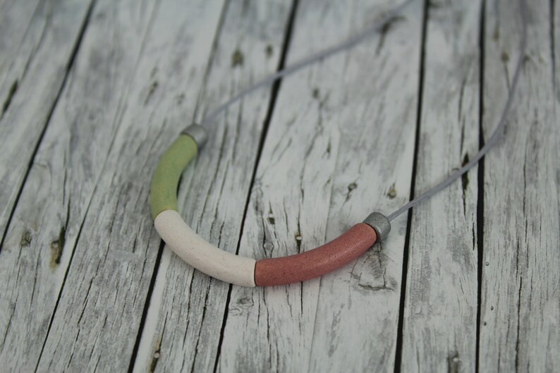 Op de afbeelding: Een ketting met een enkele, grote, veelkleurige kraal. De kraal is gemaakt van drie delen: groen, wit en roze. De ketting is gemaakt van een dun, grijs koord.