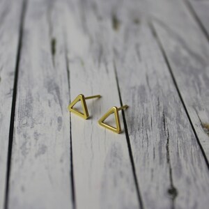 Könnte beinhalten: Zwei goldfarbene Dreiecks-Ohrstecker. Die Ohrringe sind klein, geometrisch und minimalistisch gestaltet. Sie werden auf einer hellen, verwitterten Holzoberfläche präsentiert. Die Ohrringe sind etwa 1,3 cm groß.